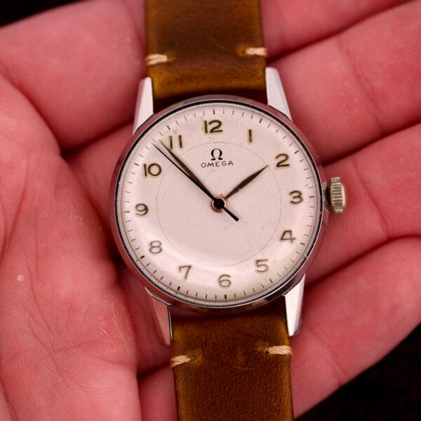 A hand holding an Omega Ref. 2496 wristwatch featuring Arabic numerals, a cream dial, brown leather strap, and stainless steel case with Cal. 30T2 SC movement.
