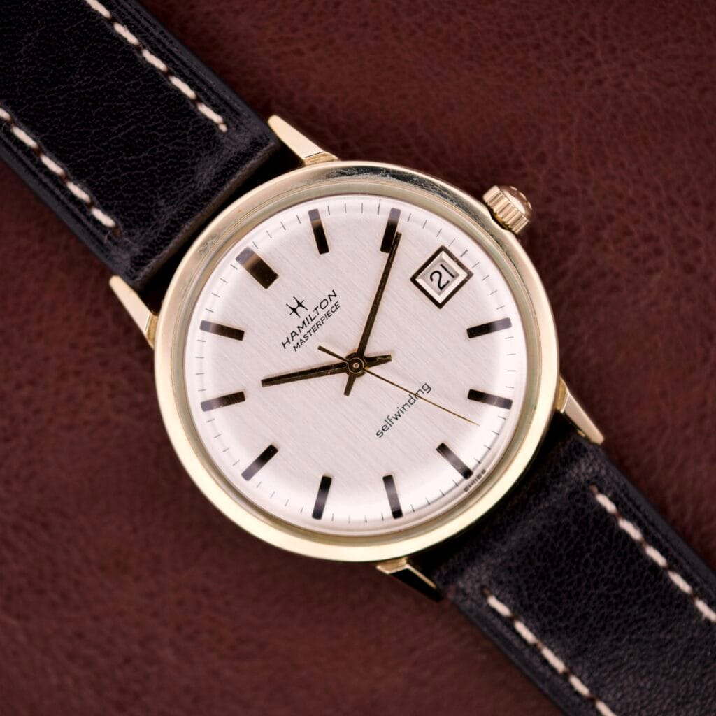 Hamilton Masterpiece Self-Winding Date wristwatch with 10K gold-filled case, white dial, black markers, date window, Cal. 822 movement, and black leather strap.