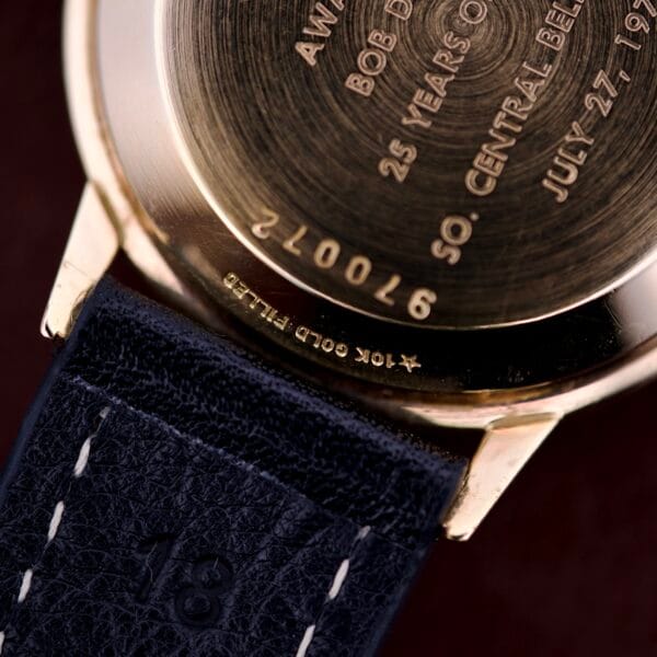 Close-up of a Hamilton Masterpiece Self-Winding Date in 10K GF Case Cal. 822, showing the engraved gold back and a dark blue leather strap with white stitching.