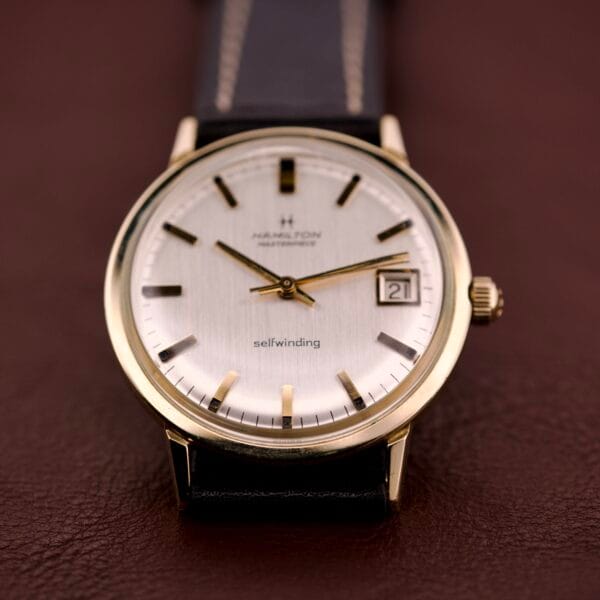 A Hamilton Masterpiece Self-Winding Date wristwatch, featuring a 10K gold-filled case (Cal. 822) and black leather strap, displayed on a brown surface.