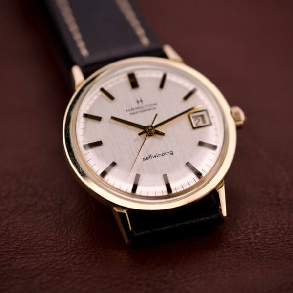 Hamilton Masterpiece Self-Winding Date wristwatch, Cal. 822, in a 10K GF case with a white dial and black leather strap, displayed on a brown surface.