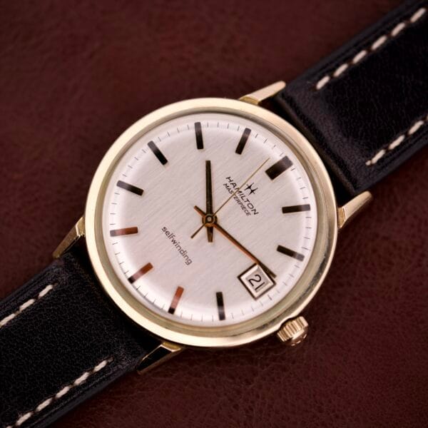 A Hamilton Masterpiece Self-Winding Date in a 10K gold-filled case (Cal. 822) with black leather strap rests on a brown surface.
