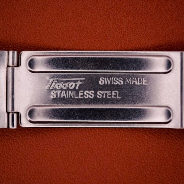 Close-up of a metal clasp engraved with Tissot Swiss Made Stainless Steel on a brown background, highlighting the elegance of the Tissot Seastar PR 516 GL Maroon Dial Ref. 46572 with day/date Cal. 794.