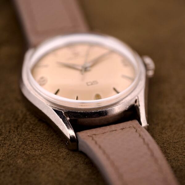A close-up of the Certina DS Ref. 346.825 1st Gen with Cal. 25-36 movement, beige dial, and brown leather strap, resting on a soft brown background beside its stainless steel case.