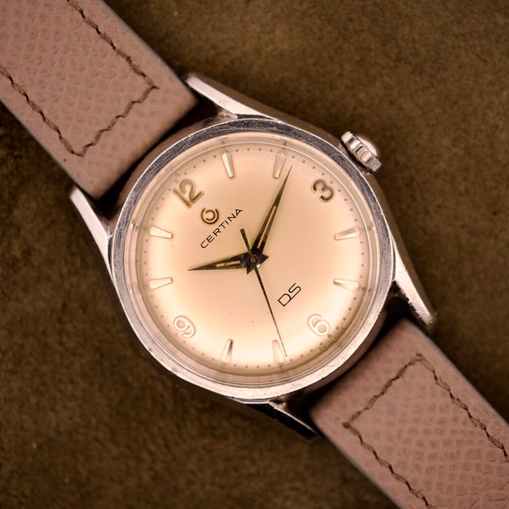 A Certina DS Ref. 346.825 1st Gen wristwatch with Cal. 25-36 movement and a beige dial in a stainless steel case with leather strap is shown resting on a brown textured surface.
