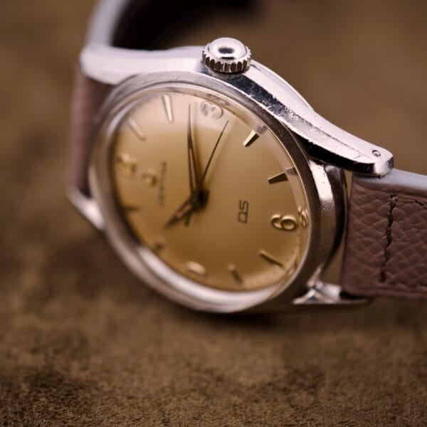 A close-up of a vintage Certina DS Ref. 346.825 1st Gen, featuring a Cal. 25-36 movement in a stainless steel case with brown leather strap, resting on a soft brown surface.