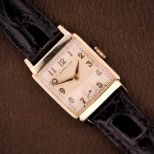 The Hamilton Tank 14k Gold Filled Case Manual Wind Cal. 982 U.S.A. wristwatch with a rectangular face and black leather strap rests on a brown textured surface.