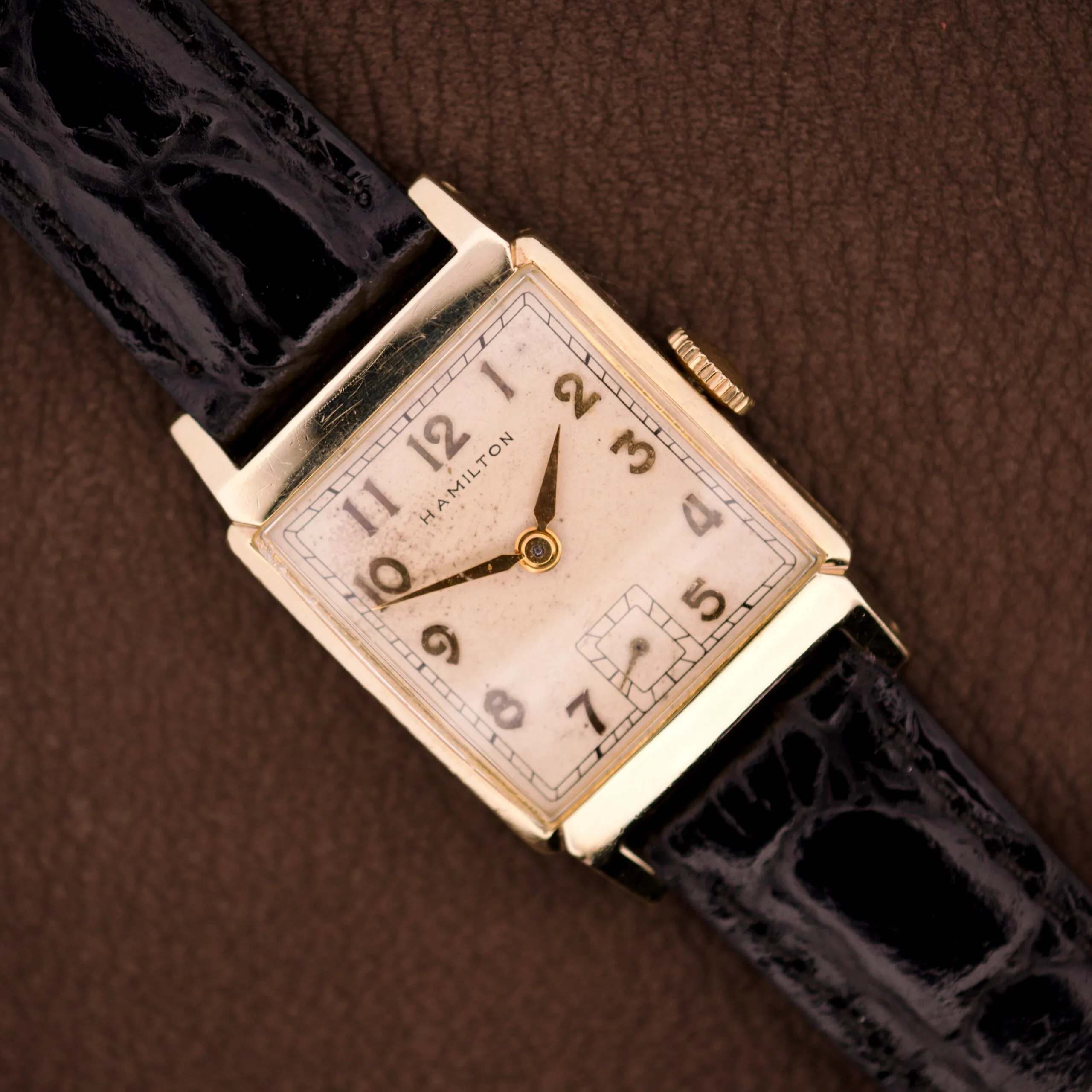 The Hamilton Tank 14k Gold Filled Case Manual Wind Cal. 982 U.S.A. wristwatch with a rectangular face and black leather strap rests on a brown textured surface.