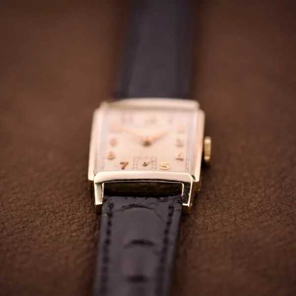 A close-up of the Hamilton Tank 14k Gold Filled Case Manual Wind Cal. 982 U.S.A. wristwatch with a black leather strap rests on a brown surface.