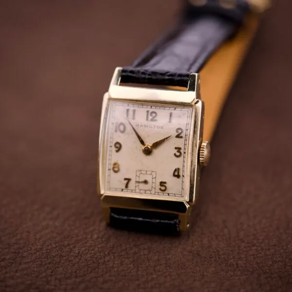 A Hamilton Tank 14k Gold Filled Case Manual Wind Cal. 982 U.S.A. wristwatch with a rectangular face and black leather strap rests on a brown surface.