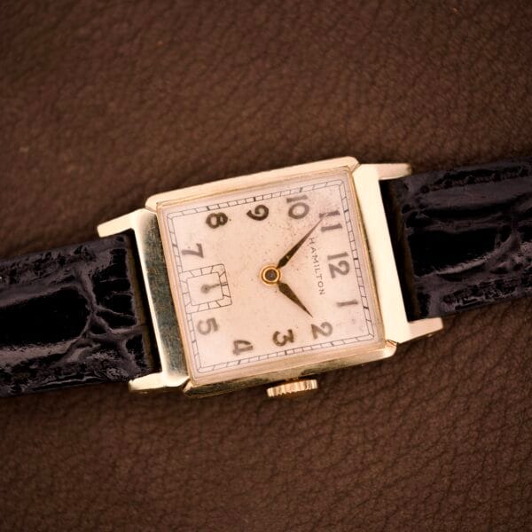 A Hamilton Tank 14k gold filled case wristwatch, featuring manual wind Cal. 982 (U.S.A.), with a black leather strap, rests on a brown surface.