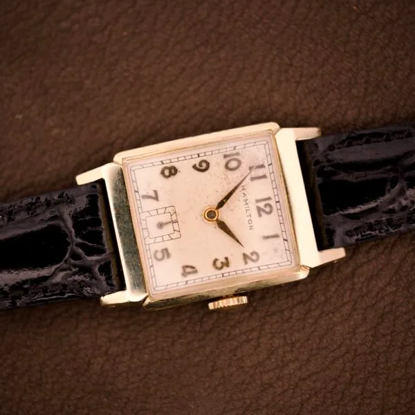 A Hamilton Tank 14k gold filled case wristwatch, featuring manual wind Cal. 982 (U.S.A.), with a black leather strap, rests on a brown surface.