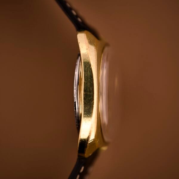 Side view of the Tissot Automatic Seastar Day/Date Cal. 2571, featuring a gold-toned case, black leather strap, and brown ombre dial set against a brown background.