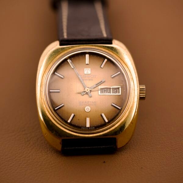 The Tissot Automatic Seastar Day/Date Cal. 2571 wristwatch with a brown ombre dial and leather band is displayed on a brown surface.