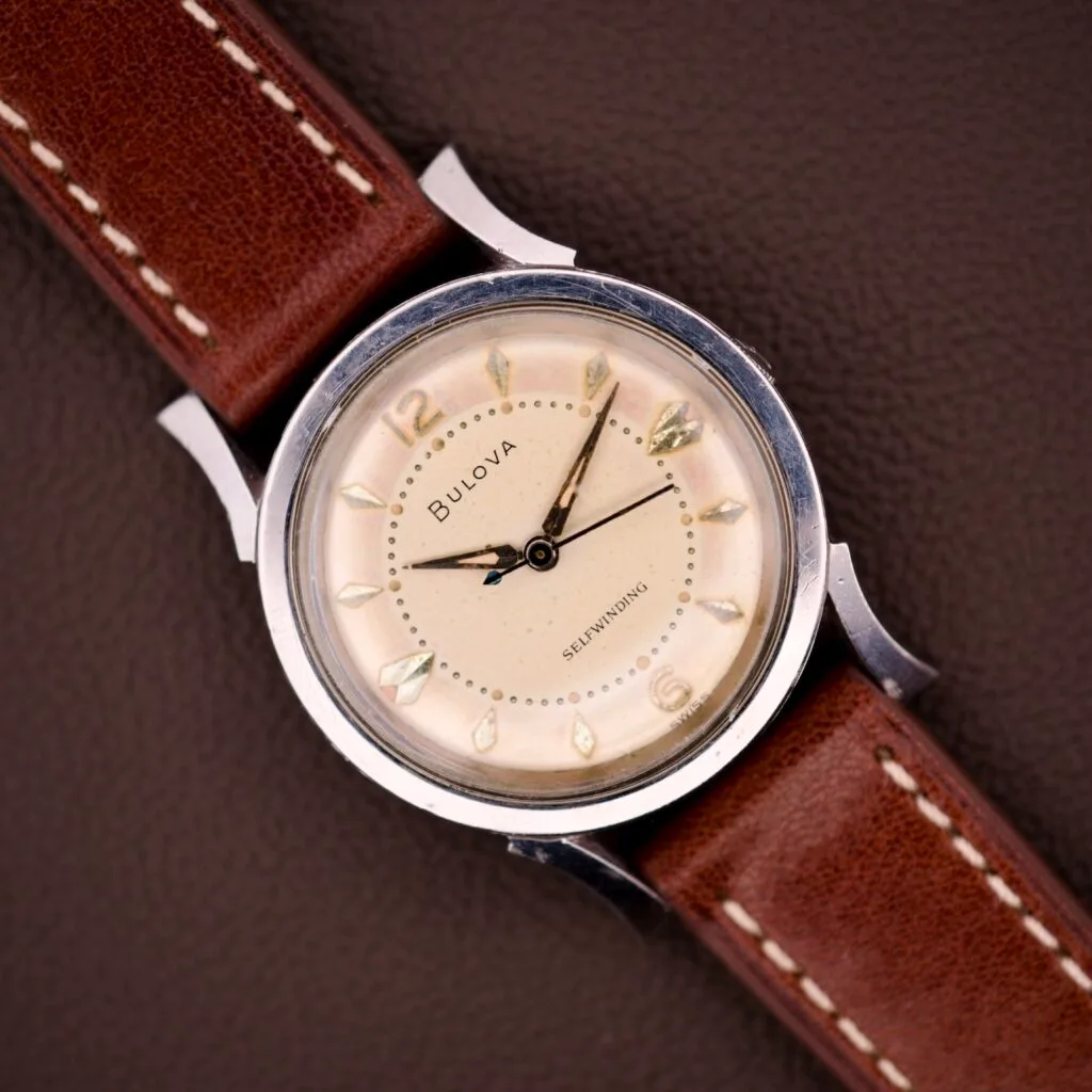 A 1959 Bulova Self-Winding Cal. 11AFAC wristwatch with a stainless steel case and fancy lugs rests on a textured brown surface.