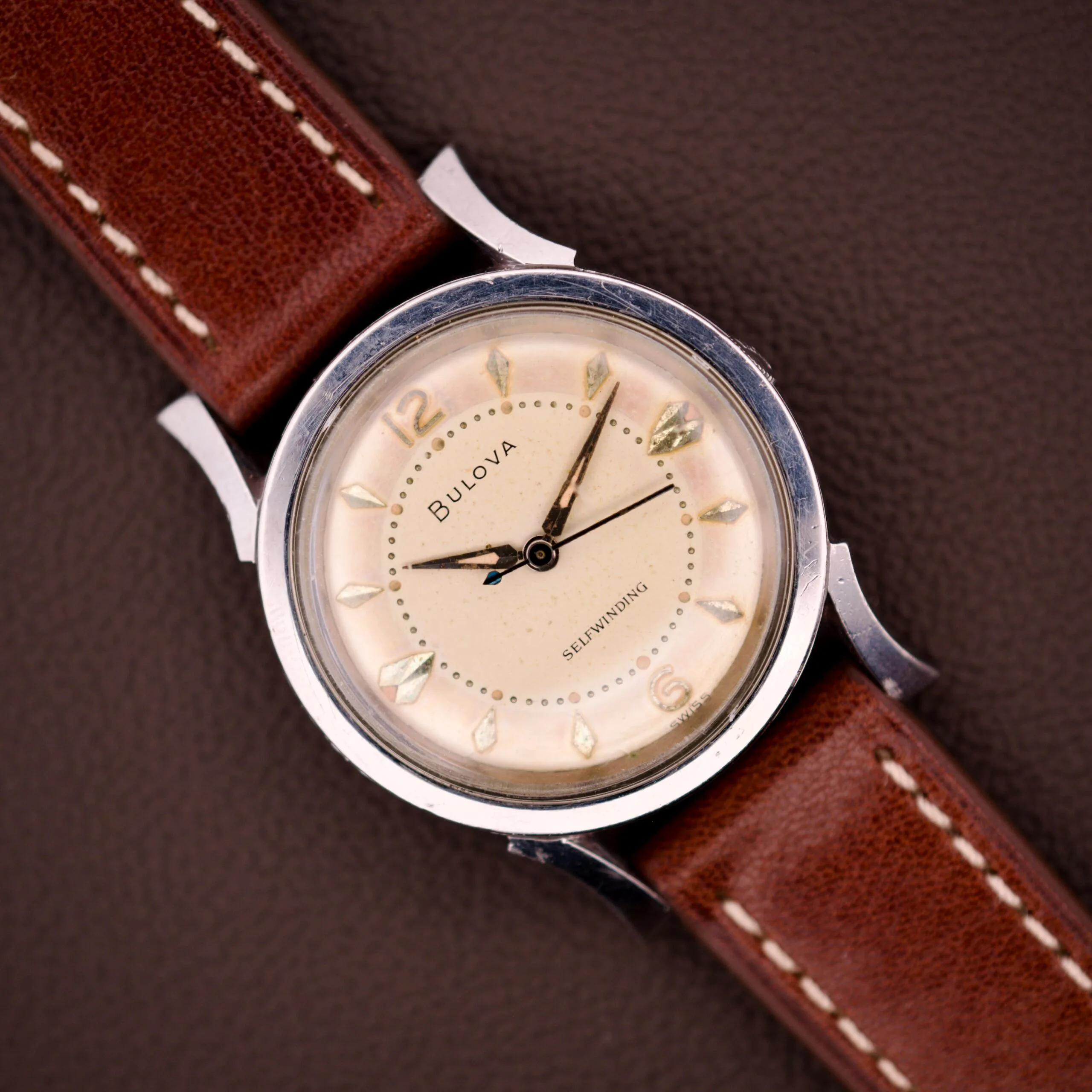 A 1959 Bulova Self-Winding Cal. 11AFAC wristwatch with a stainless steel case and fancy lugs rests on a textured brown surface.