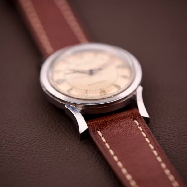 A 1959 Bulova Self-Winding Cal. 11AFAC with a stainless steel case and fancy lugs, paired with a brown leather strap, rests on a dark brown textured surface.