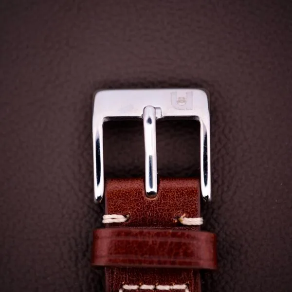 Close-up of a 1959 Bulova Self-Winding Cal. 11AFAC stainless steel buckle on a brown leather strap with white stitching, set against a dark background.