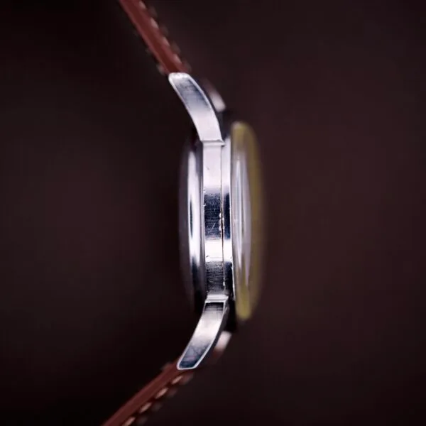 Close-up side view of the 1959 Bulova Self-Winding Cal. 11AFAC with fancy lugs, featuring a brown leather strap and a stainless steel case, set against a dark blurred background.