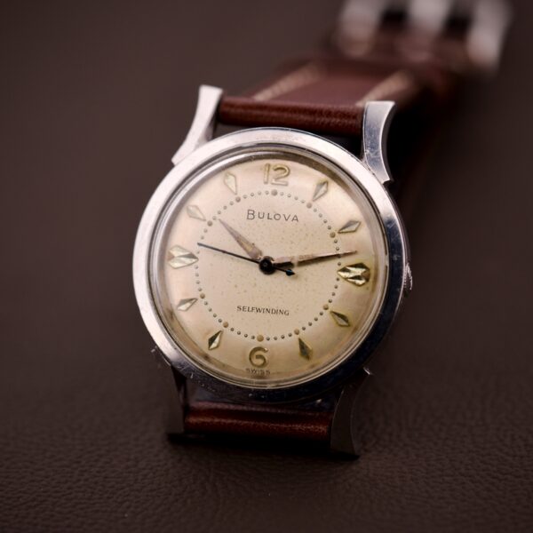 A 1959 Bulova Self-Winding Cal. 11AFAC wristwatch with fancy lugs and a stainless steel case sits on a dark brown surface.