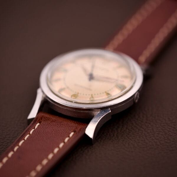 A 1959 Bulova Self-Winding Cal. 11AFAC wristwatch with a stainless steel case and fancy lugs, featuring a brown leather strap and cream dial, displayed on a brown textured surface.