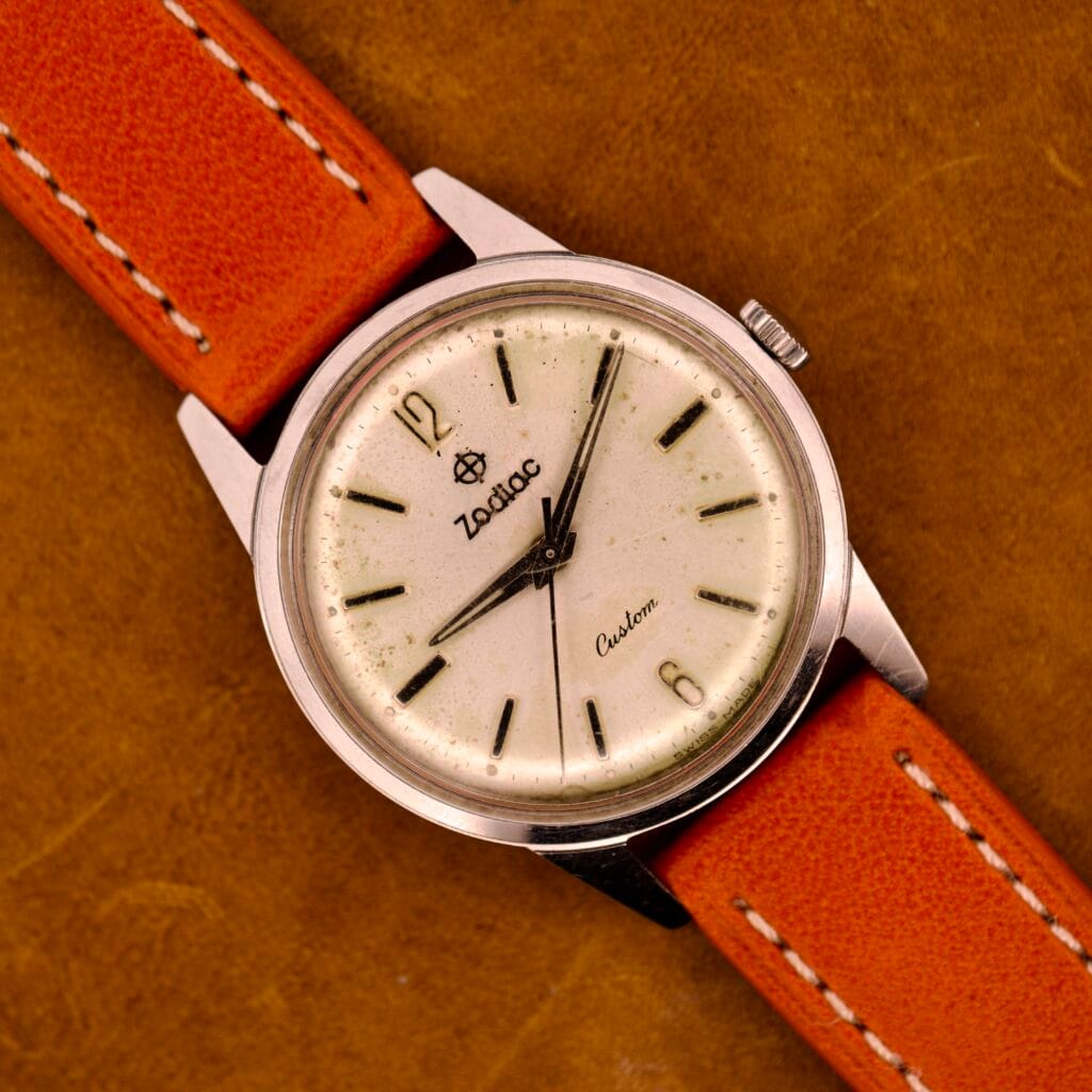 Zodiac Hermetic Custom Manual Wind wristwatch in stainless steel case, featuring a cream dial with black markers and an orange leather strap, displayed on a brown surface.