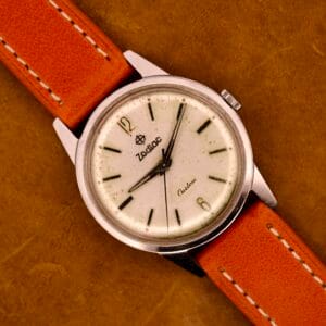 Zodiac Hermetic Custom Manual Wind wristwatch in stainless steel case, featuring a cream dial with black markers and an orange leather strap, displayed on a brown surface.