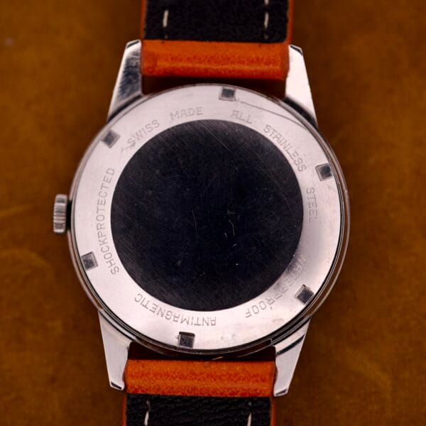 Back of a Zodiac Hermetic Custom Manual Wind in stainless steel case with orange and black strap, shown on a brown surface.