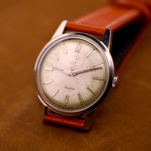 A Zodiac Hermetic Custom Manual Wind wristwatch with a stainless steel case and leather strap rests on a brown surface.