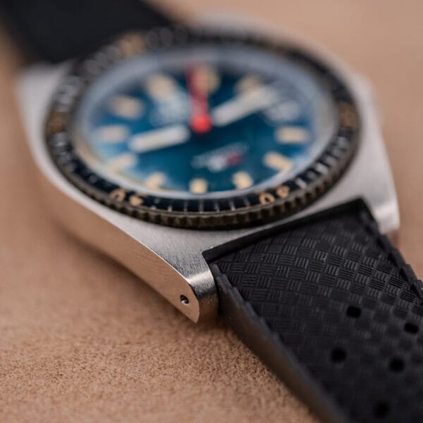 Close-up of a Tissot PR-516 "Roger Moore" Bakelite Bezel Ref. 44622 wristwatch with a black textured strap and blue dial, shown on a tan surface.