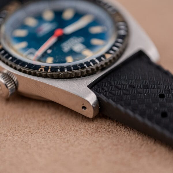 Close-up of a Tissot PR-516 "Roger Moore" Balkelite Bezel Ref. 44622 with a black textured strap and blue dial, resting on tan—evoking the timeless style associated with Roger Moore.