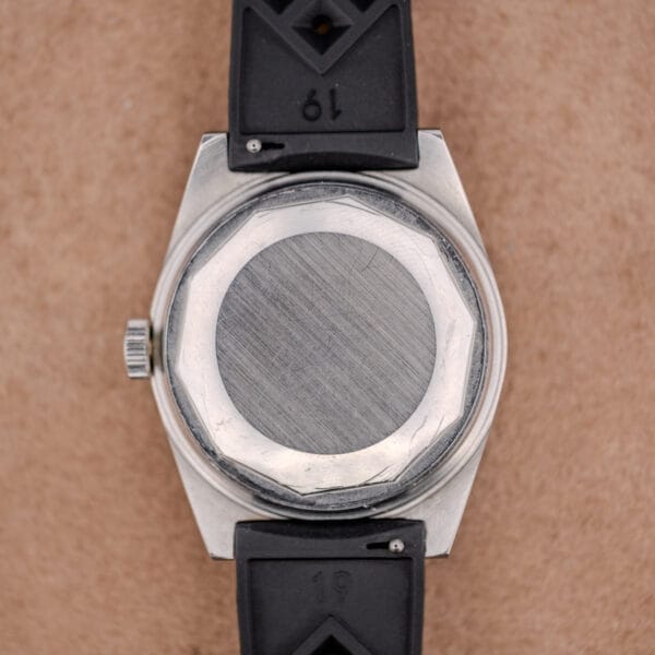 The back of a Tissot PR-516 "Roger Moore" Balkelite Bezel Ref. 44622 wristwatch with a black strap, displayed against a tan background.