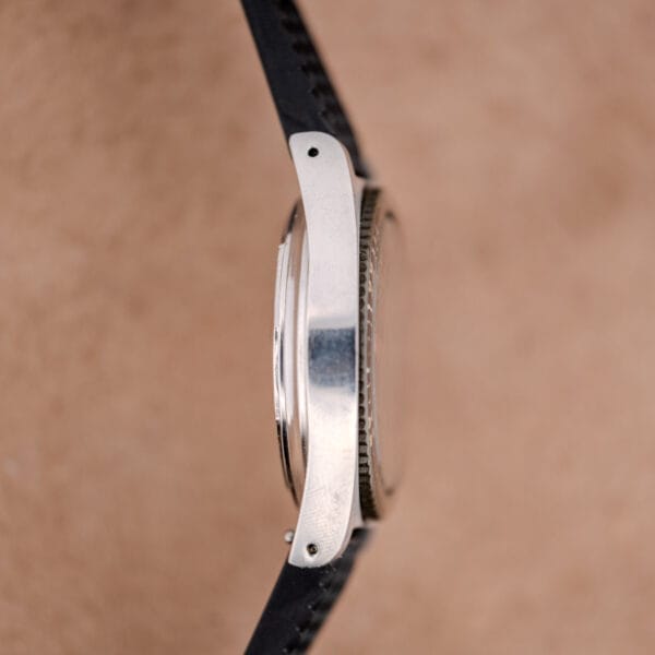 Side view of a silver Tissot PR-516 "Roger Moore" Balkelite Bezel Ref. 44622 case with black strap, set against a beige background.
