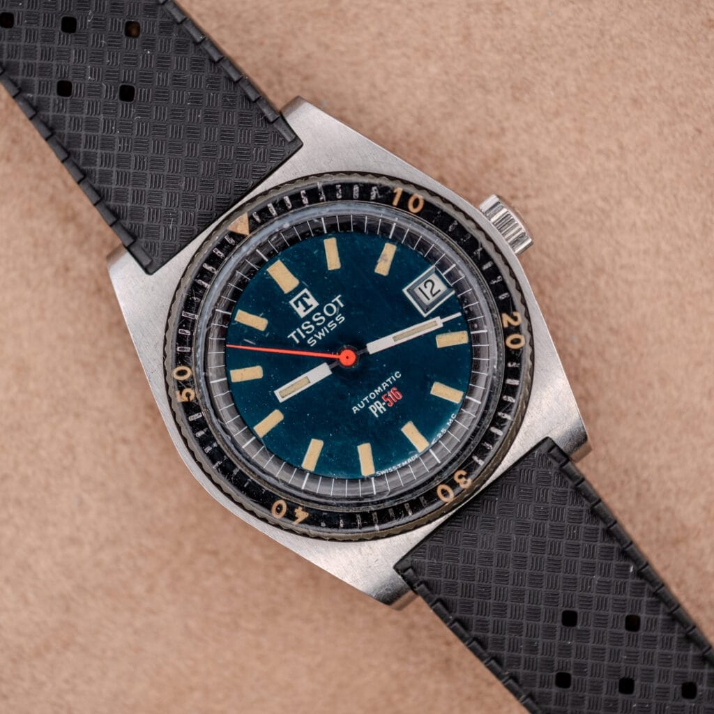 A Tissot PR-516 "Roger Moore" Balkelite Bezel Ref. 44622 wristwatch with a blue dial, date window, and black textured strap rests on a tan surface, reflecting timeless style.