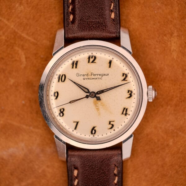 Girard Perregaux Gyromatic in stainless steel with Arabic numerals dial, shown on a brown leather strap and elegantly placed on a tan leather surface.
