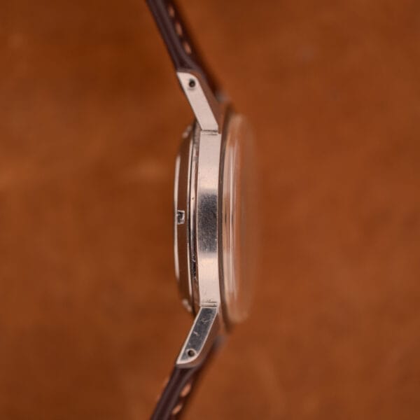 Side view of a Girard Perregaux Gyromatic in stainless steel with Arabic numerals dial and brown leather strap, displayed against a matching brown background.