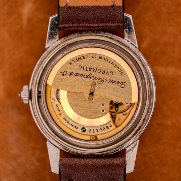Close-up of a vintage Girard Perregaux Gyromatic in stainless steel with Arabic numerals dial, automatic movement, and brown leather strap on a tan background.