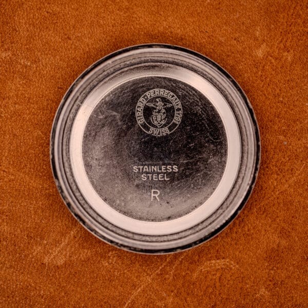 A Girard Perregaux Gyromatic in Stainless Steel with Arabic numerals dial is displayed on a brown textured surface, its case back inscribed with GIRARD PERREGAUX SWISS.