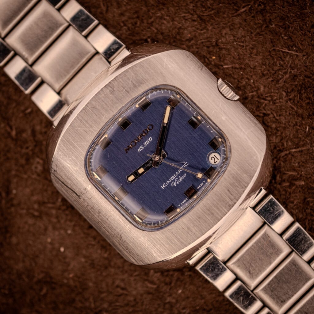 A Movado HS 360 Kingmatic Video Blue Dial Ref. 409-705 Cal. 408 wristwatch with a blue square dial and silver metal band, displayed on a brown surface.