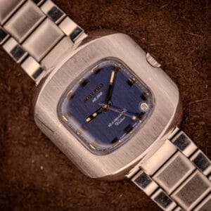 A Movado HS 360 Kingmatic Video Blue Dial Ref. 409-705 Cal. 408 wristwatch with a blue square dial and silver metal band, displayed on a brown surface.