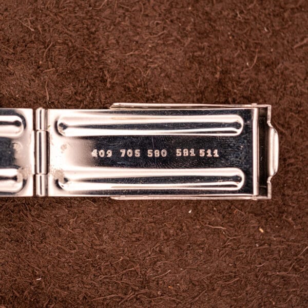 A metal clasp engraved with 409 705 580 581 511, likely from a Movado HS 360 Kingmatic Video Blue Dial Ref. 409-705 Cal. 408, rests on a brown textured surface.