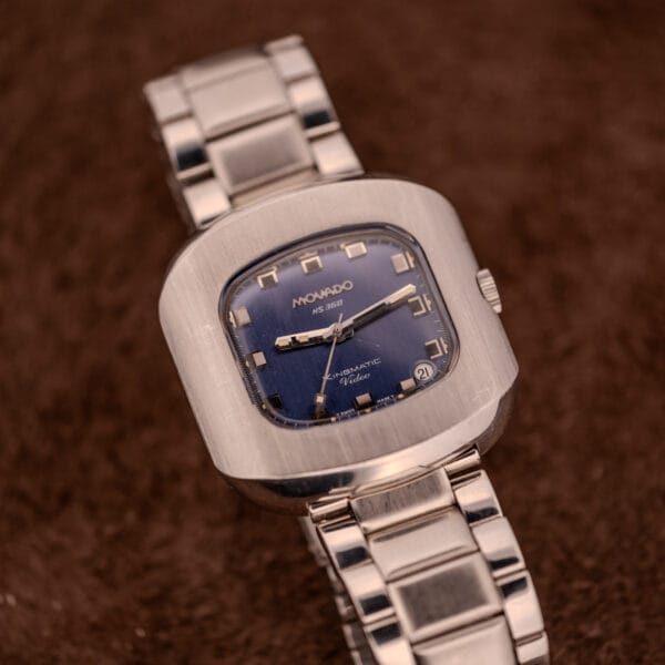 A Movado HS 360 Kingmatic Video Blue Dial Ref. 409-705 Cal. 408 wristwatch with a silver case and blue square dial is displayed on a brown textured surface.