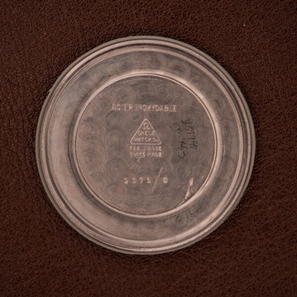 Stainless steel case back from an Omega WWII Era Military "US ARMY" Ref. 2179 Cal. 30T2 SC watch, showing Omega markings and handwritten notes, displayed on a brown leather surface.