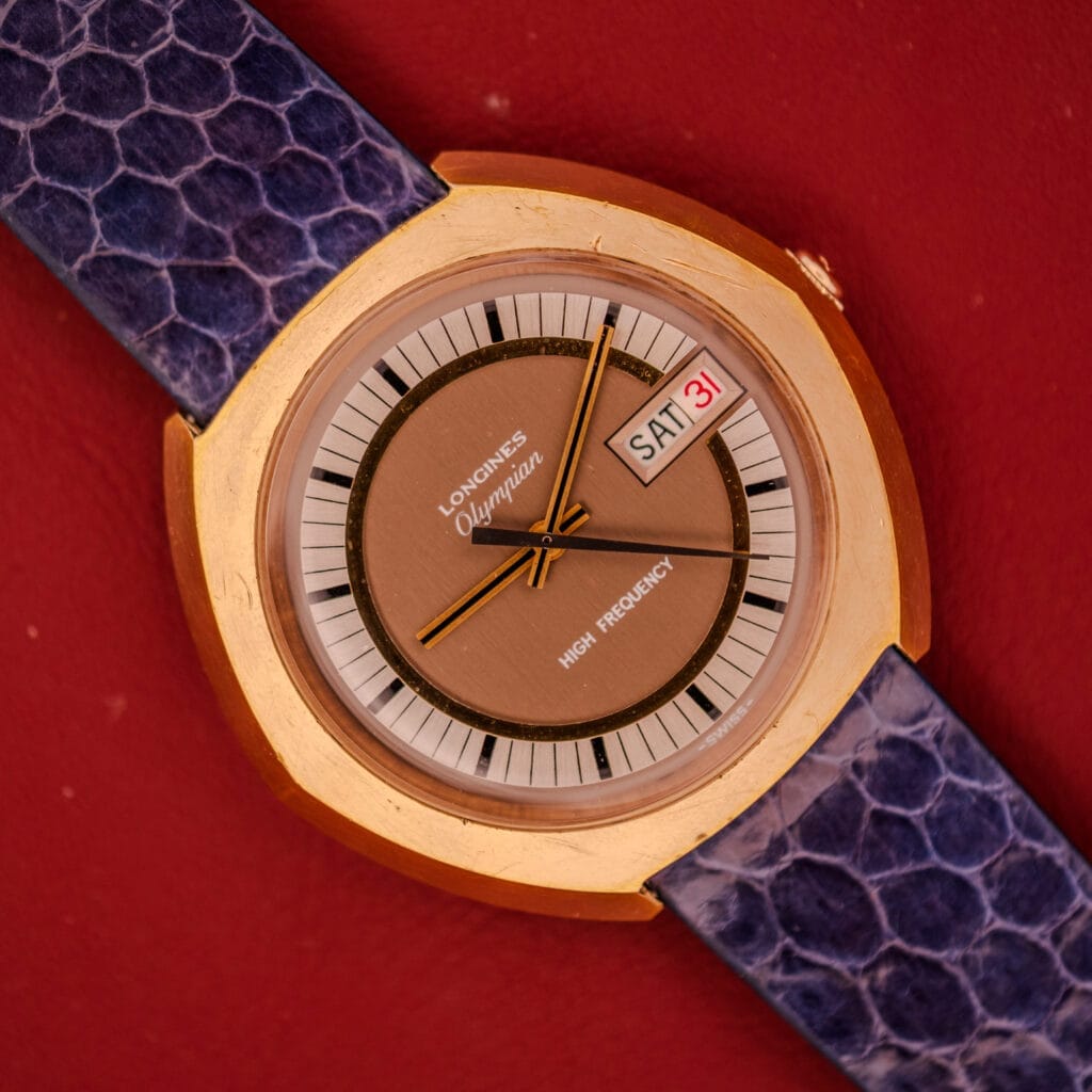A vintage Longines Olympian High Frequency Ref. 3141 watch with Cal. 6972 movement, gold case, purple strap, and day-date display on a red background.