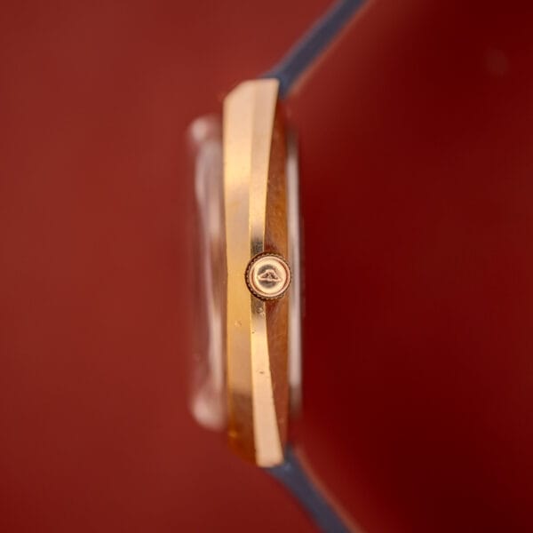 Close-up side view of a Longines Olympian High Frequency Ref. 3141 gold case and crown, featuring the Cal. 6972 movement, set against a blurred red background.