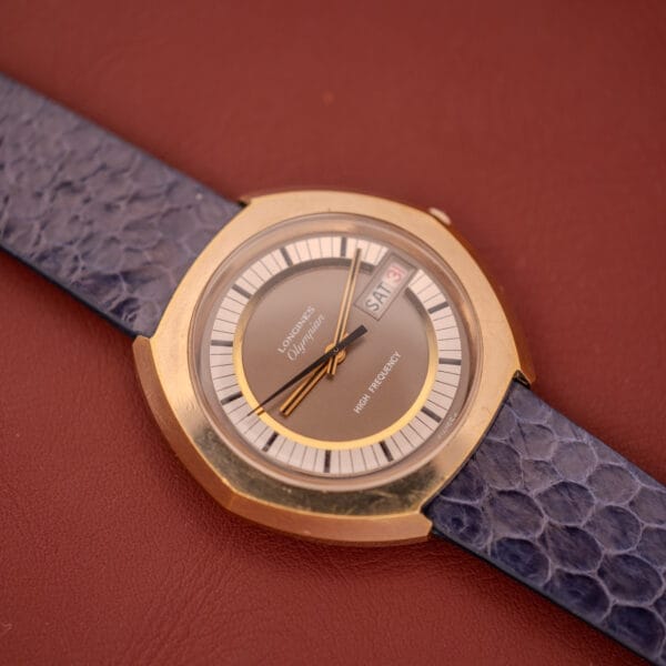 A Longines Olympian High Frequency Ref. 3141 Cal. 6972 watch with a blue strap, gold case, and day-date display rests on a brown surface.