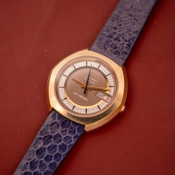 A Longines Olympian High Frequency Ref. 3141 Cal. 6972 wristwatch with a gold case and blue strap displays day and date, shown on a red leather surface.