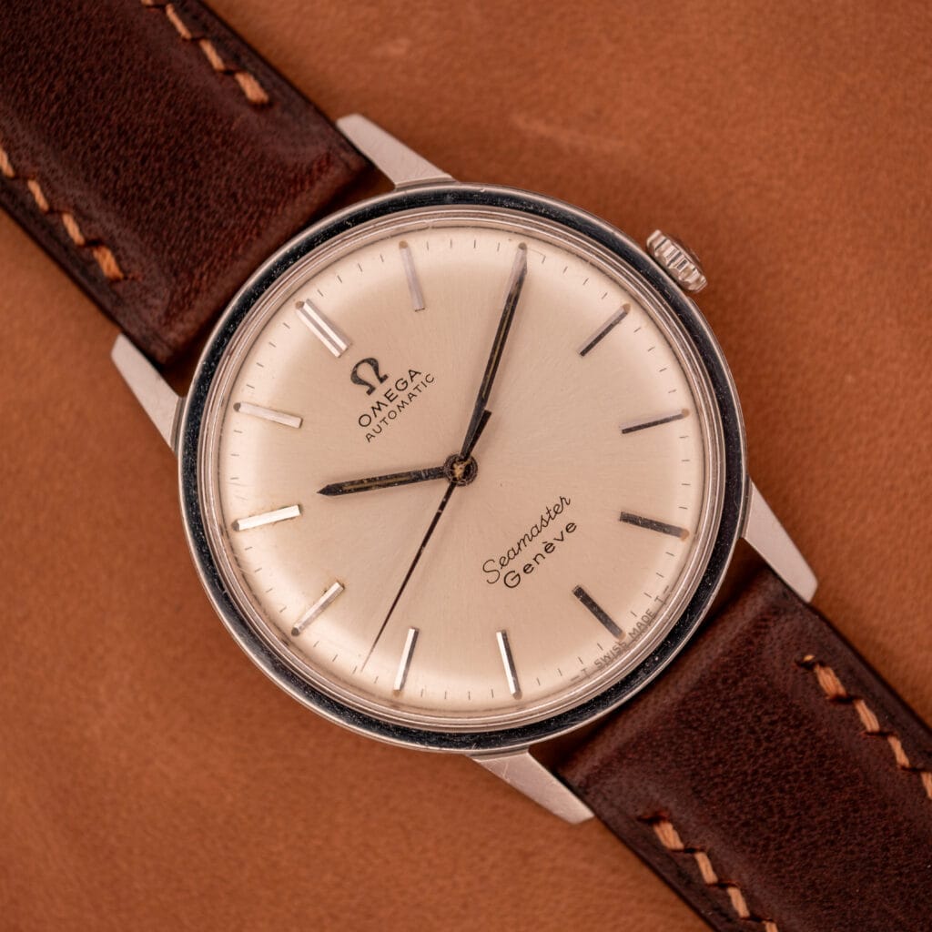 An Omega Automatic Seamaster Geneve Double Signed Dial Ref. 165.002 with a brown leather strap rests on a tan surface.