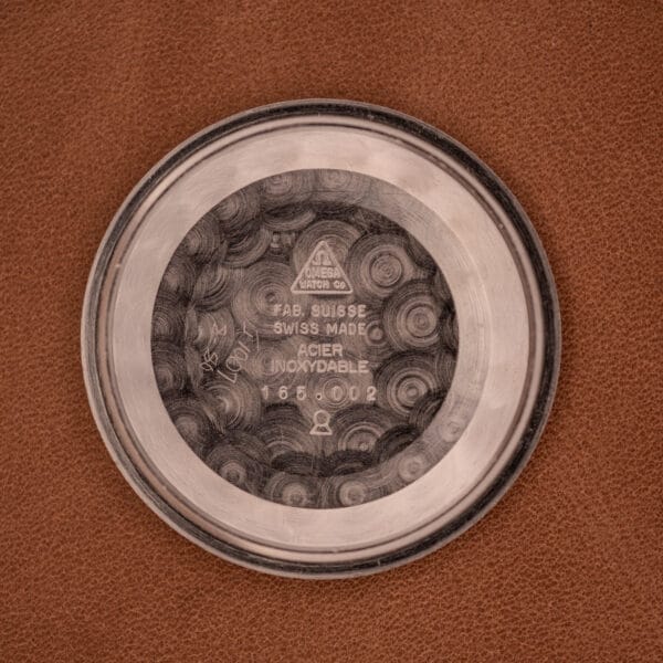 The Omega Automatic Seamaster Geneve Double Signed Dial Ref. 165.002 features a round metal case back with engraved Seamaster branding, Swiss-made marks, and hints at its automatic movement, displayed on a brown surface.