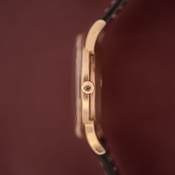 Side view of a Girard Perregaux Gyromatic wristwatch with a 10k gold filled case and cream dial, paired with a brown leather strap, set against a burgundy background.